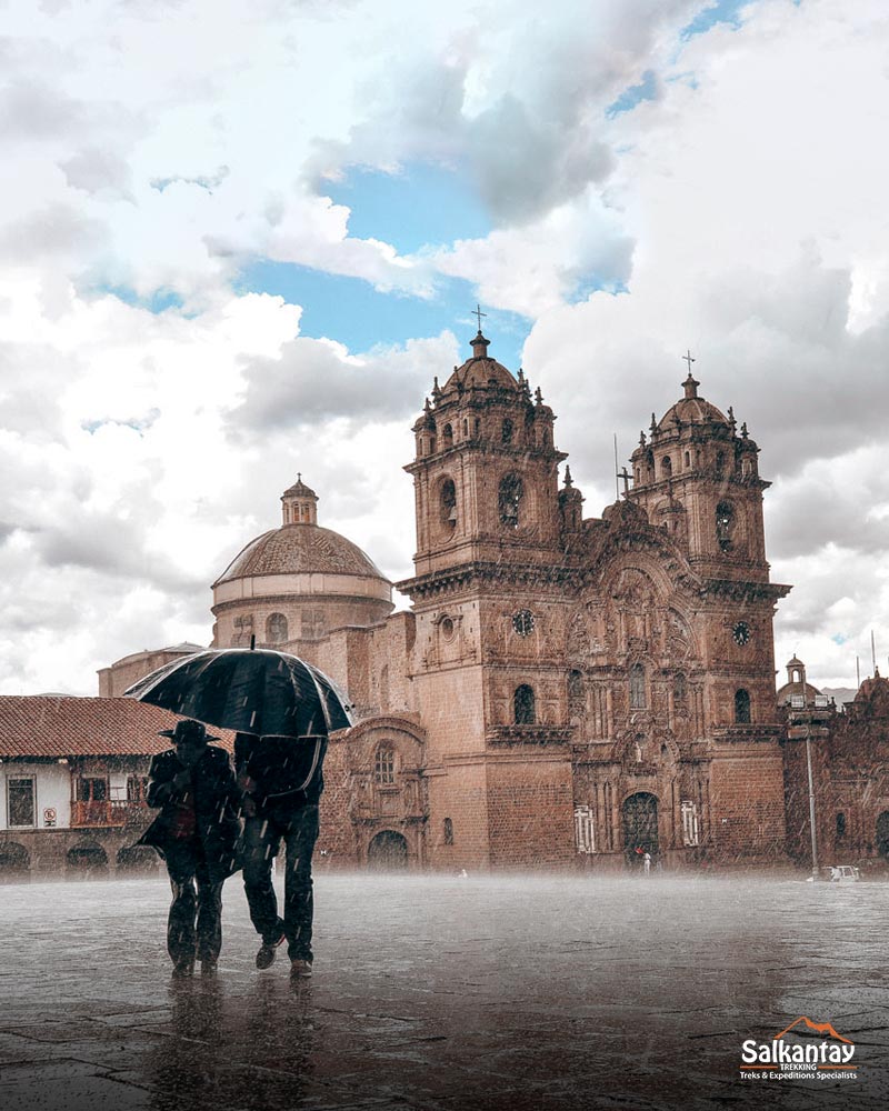 Dois turistas com guarda-chuvas na praça principal de Cusco em meio à chuva torrencial.